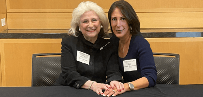 Two women sitting at a table and smiling warmly while holding hands. One has white hair and is wearing a black outfit with a name tag that reads “Fordham Law.” The other has long dark hair, is dressed in a navy top, and is also wearing a name tag. They appear to be at a formal or professional event.